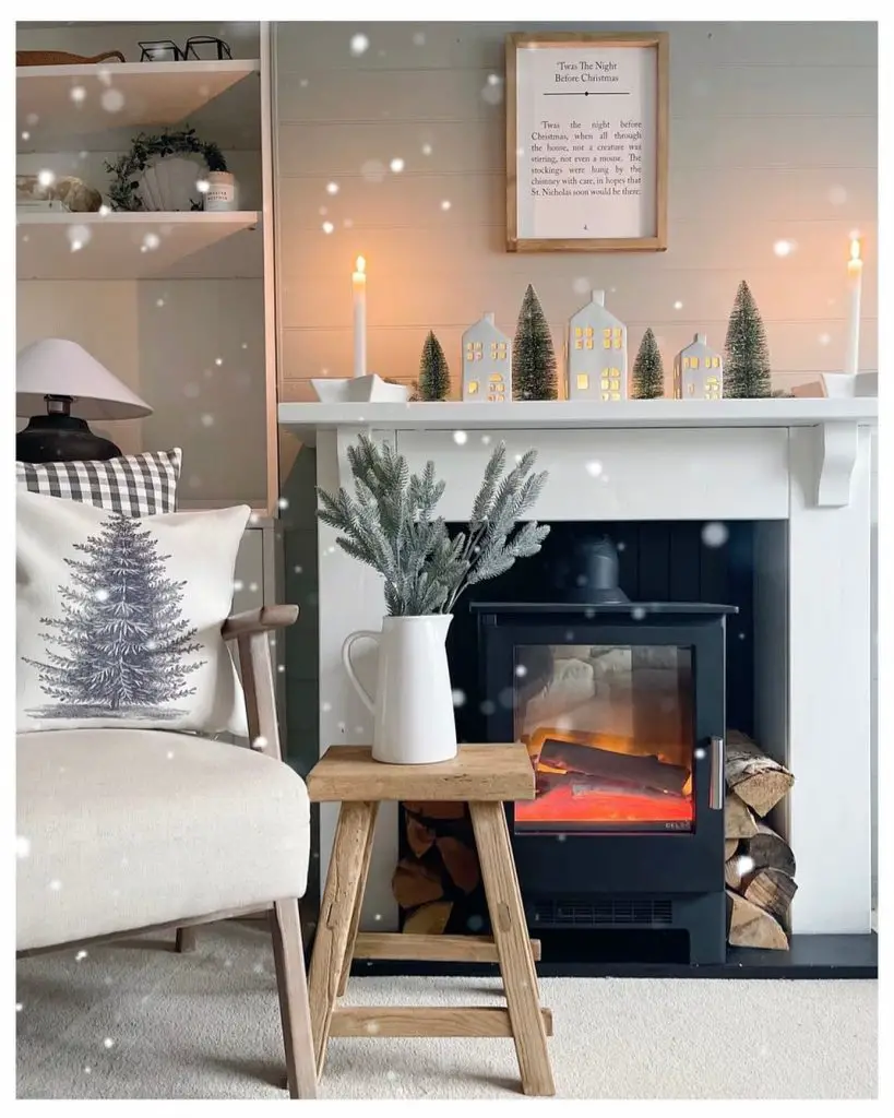 A cozy living room features a lit electric fireplace, a wooden stool with a vase, small Christmas trees on the mantel, and a beige chair with a tree-patterned cushion. Snowflakes appear to be falling, perfectly illustrating how to embrace the season and transition your home decor from fall to winter.