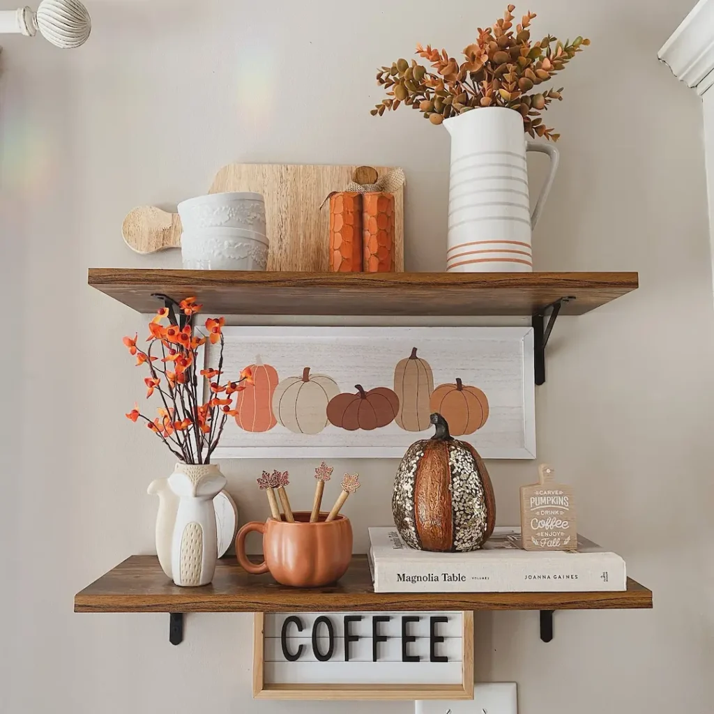 Two wooden wall shelves display autumn-themed decor, including vases with orange leaves, a pumpkin ornament, jars, and a framed pumpkin illustration—capturing the spirit of seasonal transitions. A “COFFEE” sign sits below the shelves.