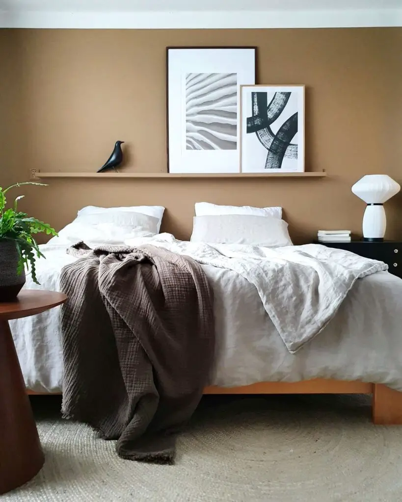 A neatly made bed with beige linens is set against a brown wall adorned with two framed artworks, showcasing how to incorporate meaningful artifacts in your Japandi bedroom. To the left is a wooden side table with a potted plant, and to the right is a black nightstand with a lamp.