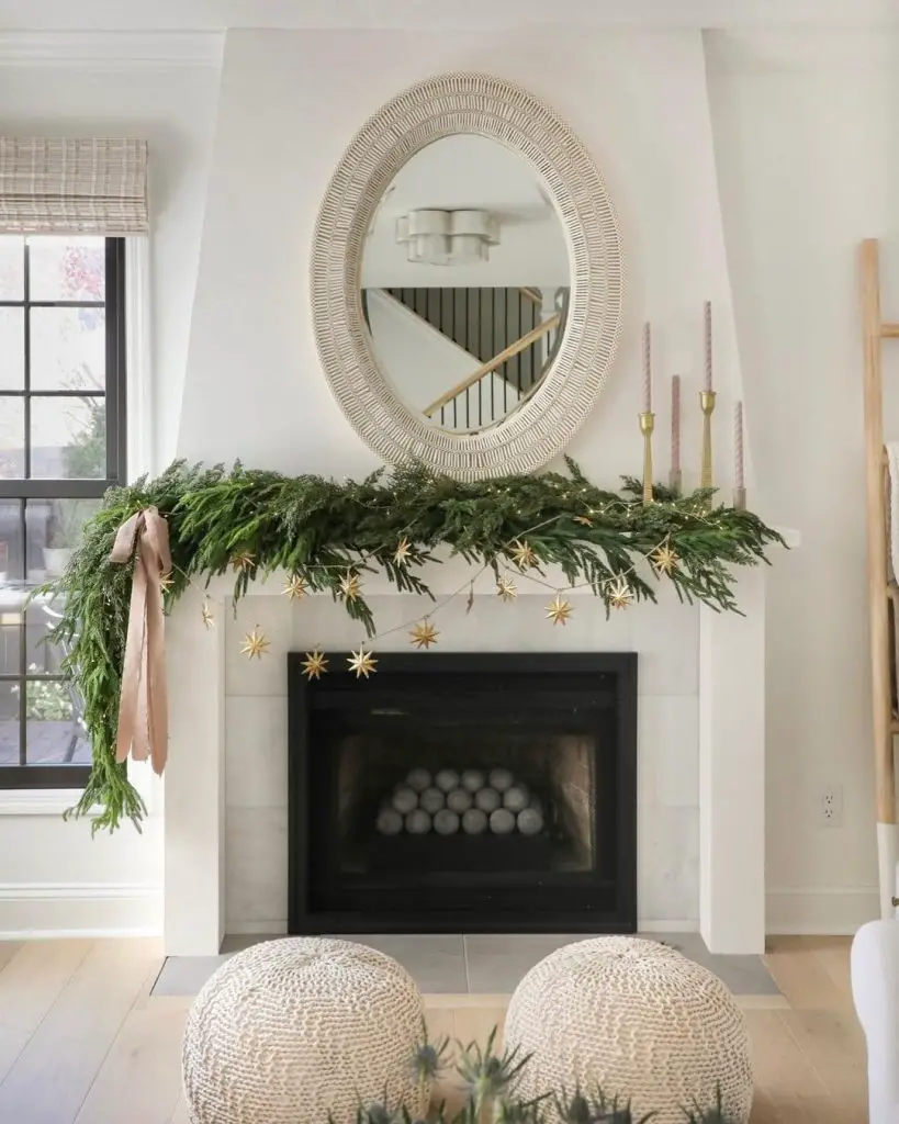 A fireplace decorated with greenery, gold star ornaments, and pink candles stands below a round wicker-framed mirror in a bright living room with light wooden floors and a window with checkered curtains, embracing minimalism: how to create a chic winter decor scheme effortlessly.