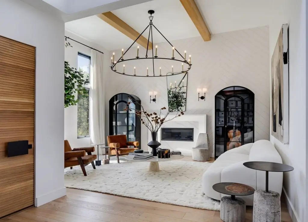 A modern living room with a large black chandelier, white walls, wooden beams, a white plush rug, a white sofa, wooden chairs. Art pieces adorn the wall, and plants bask near the windows—embodying a Modern Rustic Style: The Trendy Twist on Timeless Comfort.
