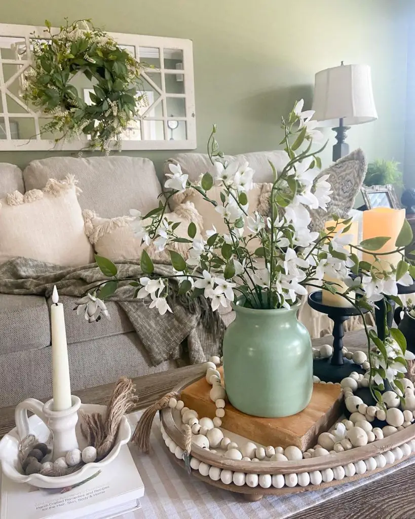 A cozy living room features a beige sofa with pillows, a green vase with white flowers on a decorative tray on the coffee table, and a wreath above a window frame on the wall—perfect elements for seamless seasonal transitions.