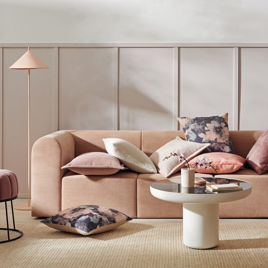 A modern living room with a beige couch adorned with various pillows in pastel colors. A round coffee table holds a vase with flowers and books, showcasing The Power of Texture: How to Create Visual Depth in Your Living Room. A floor lamp stands in the background.