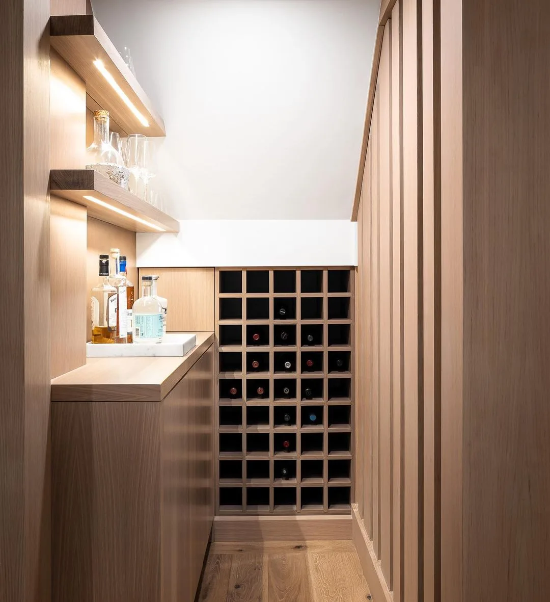A contemporary wine cellar with wooden shelving, several bottles of various spirits on a counter, and a grid-style wine rack holding multiple bottles of wine. This setup is one of the 10 creative under stairs storage ideas to optimize your space, featuring soft and warm lighting throughout.