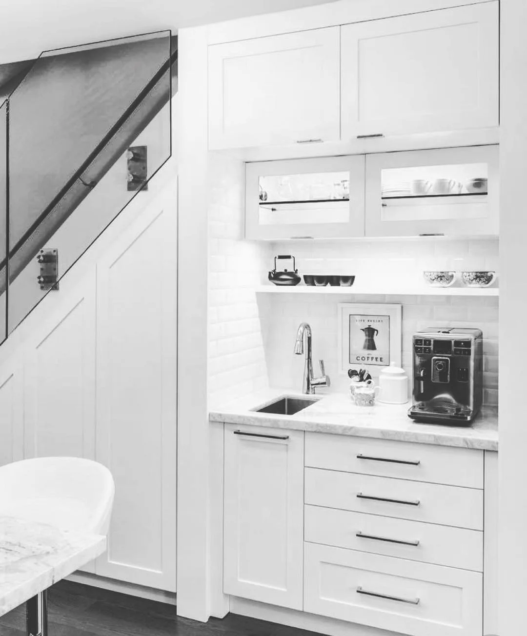 A monochromatic kitchen features a small sink, a coffee machine on a marble countertop, overhead cabinets, and drawers below. Near the staircase are minimalist, clean white cabinetry inspired by 10 creative under stairs storage ideas to optimize your space.