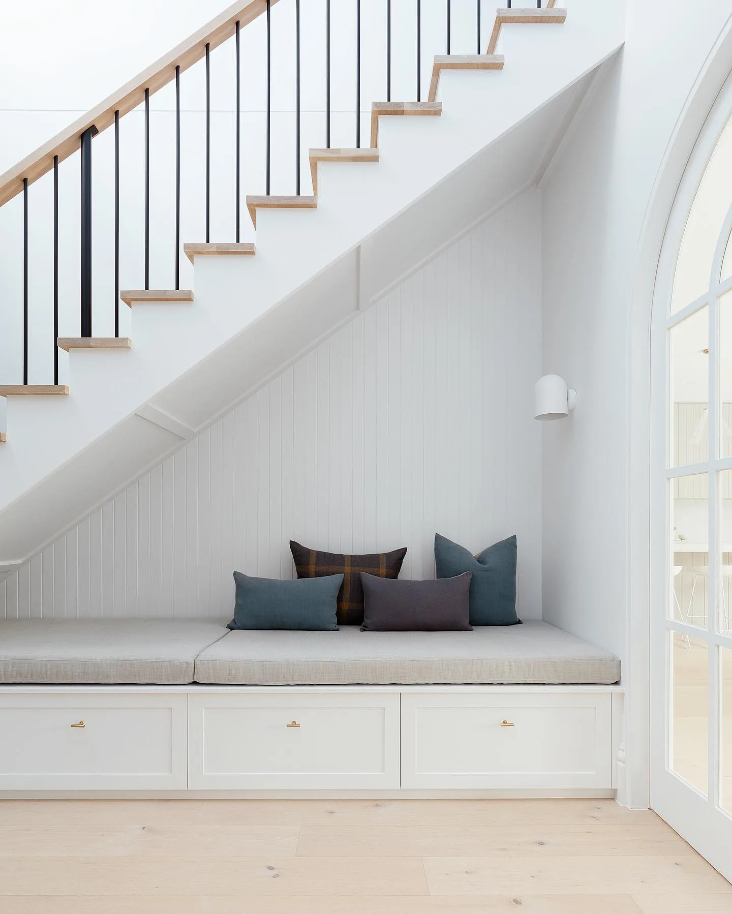 A small nook with a cushioned bench and pillows is tucked under a staircase. The space has white walls, a wooden floor, and a black metal stair railing. An arched doorway is visible on the right, showcasing one of the 10 Creative Under Stairs Storage Ideas to Optimize Your Space.