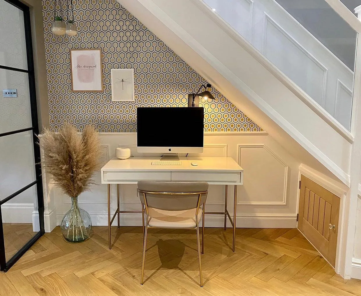 A compact workspace with a desk, chair, and computer set up under a staircase. The area features a wallpapered wall, framed pictures, a glass vase with pampas grass, and a small cabinet – an excellent example of 10 Creative Under Stairs Storage Ideas to Optimize Your Space.