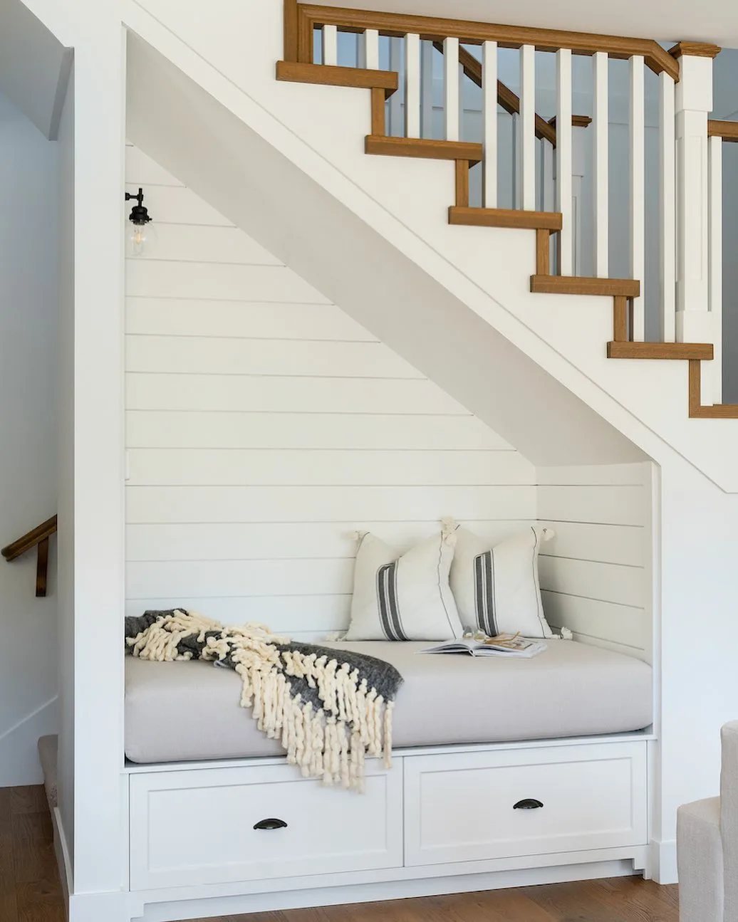 A built-in reading nook with a cushioned seat, two pillows, a throw blanket, and a book under a staircase with white shiplap walls and wooden accents is one of the 10 creative under-stairs storage ideas to optimize your space.