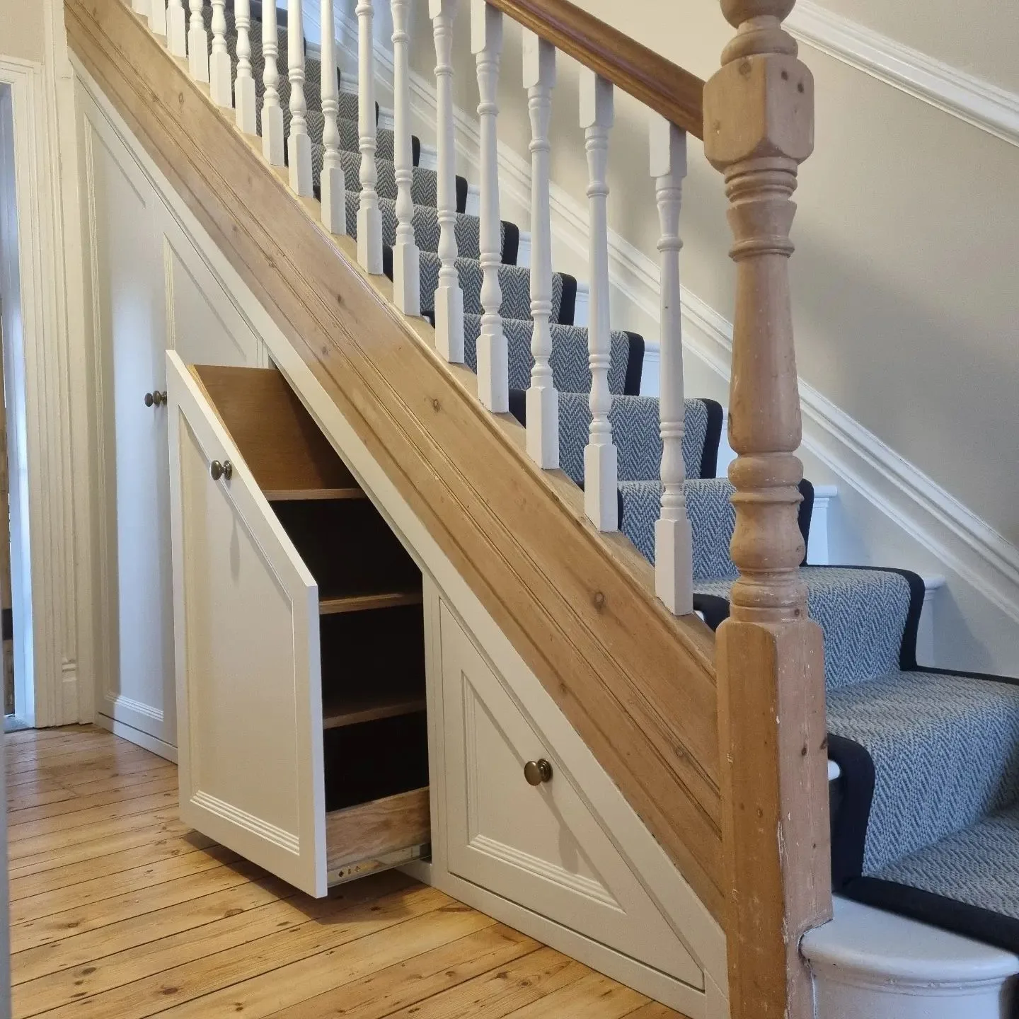 A wooden staircase with carpeted steps and white balusters features a pull-out storage drawer under the stairs, embodying one of the 10 creative under stairs storage ideas to optimize your space.