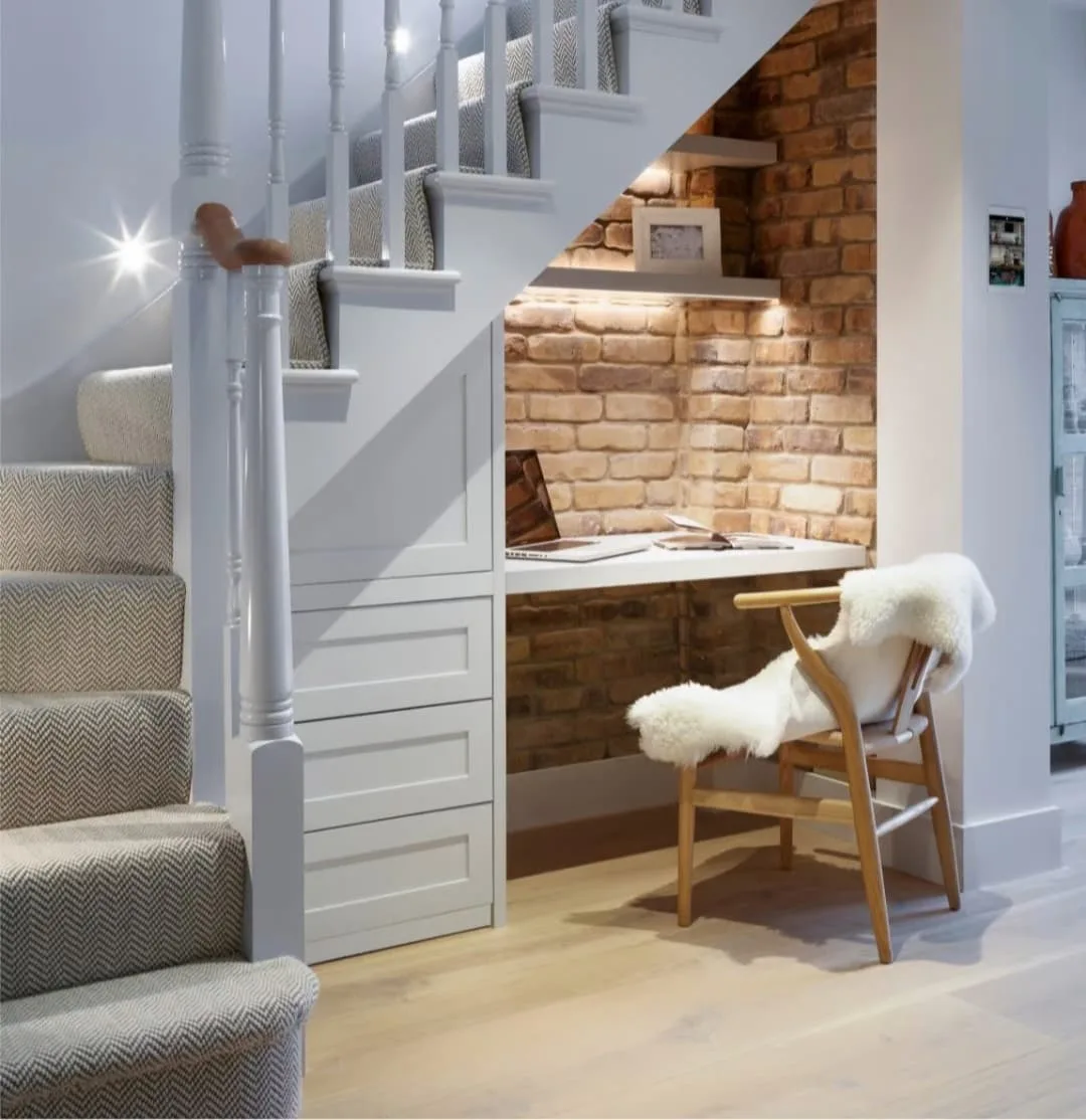 A cozy workspace under a staircase with a wooden desk, shelves, chair with a white throw, and a brick wall background is one of the 10 creative under stairs storage ideas to optimize your space.