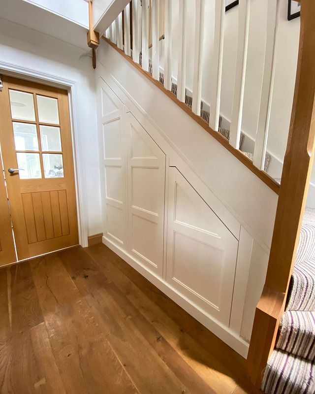 A hallway features a wooden door with glass panels on the left and built-in storage cabinets under a staircase with white railings and wood accents. The floor is made of hardwood, creating an ideal spot for an under stairs pantry: a dream storage solution for small kitchens.