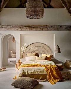 A bedroom features a bed with neutral and yellow bedding, a wicker headboard, a woven pendant light, and rustic wooden beams. Embracing The Beauty of Imperfection: How Wabi-Sabi Enhances Japandi Bedroom Decor, the room includes a side table with a vase and various woven baskets.