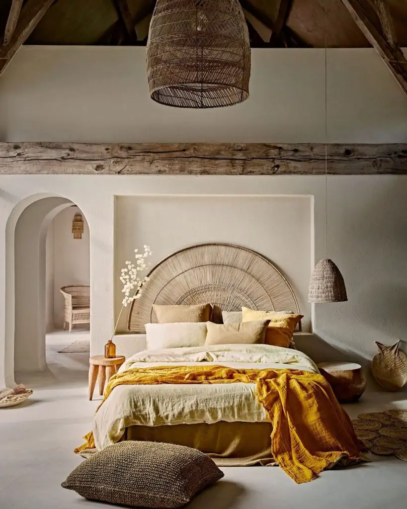 A bedroom features a bed with neutral and yellow bedding, a wicker headboard, a woven pendant light, and rustic wooden beams. Embracing The Beauty of Imperfection: How Wabi-Sabi Enhances Japandi Bedroom Decor, the room includes a side table with a vase and various woven baskets.