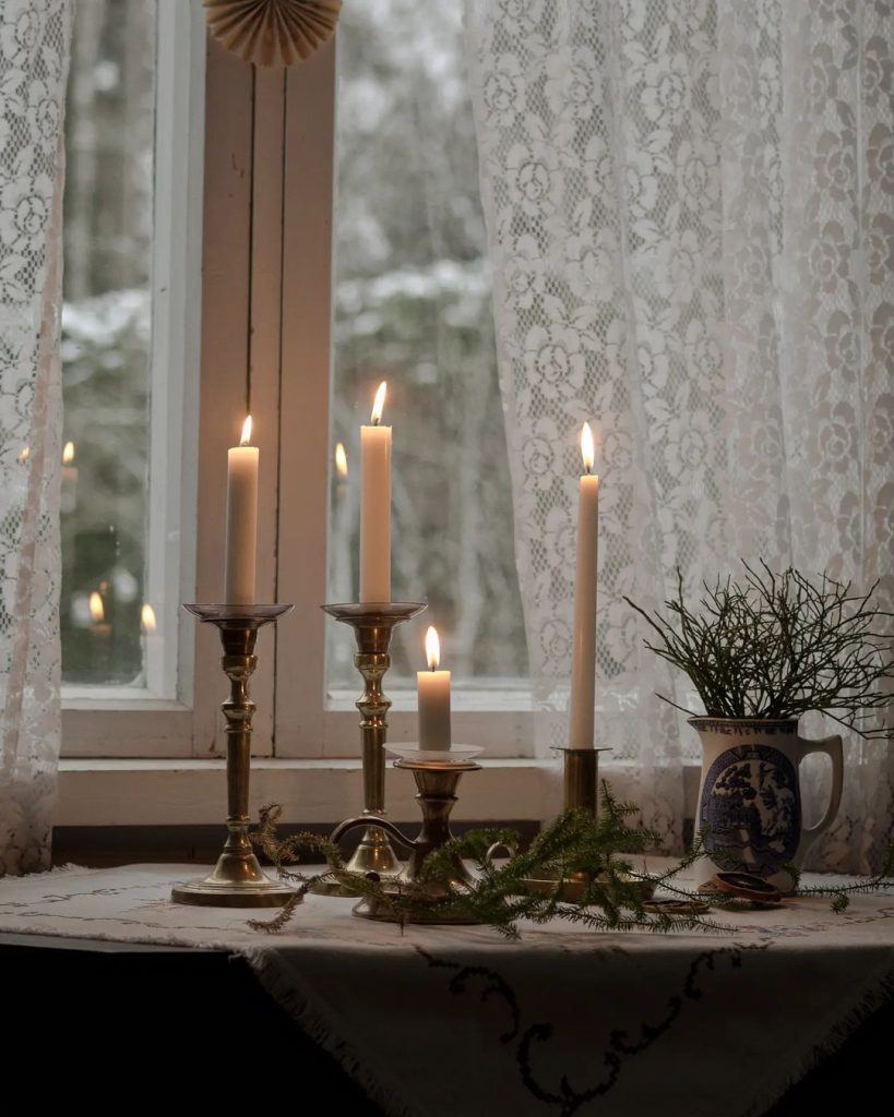 A windowsill with four lit candles in brass holders, a mug with a small plant, lace curtains, and a snowy scene outside—Winter Hygge Essentials at their finest for a warm and inviting home.