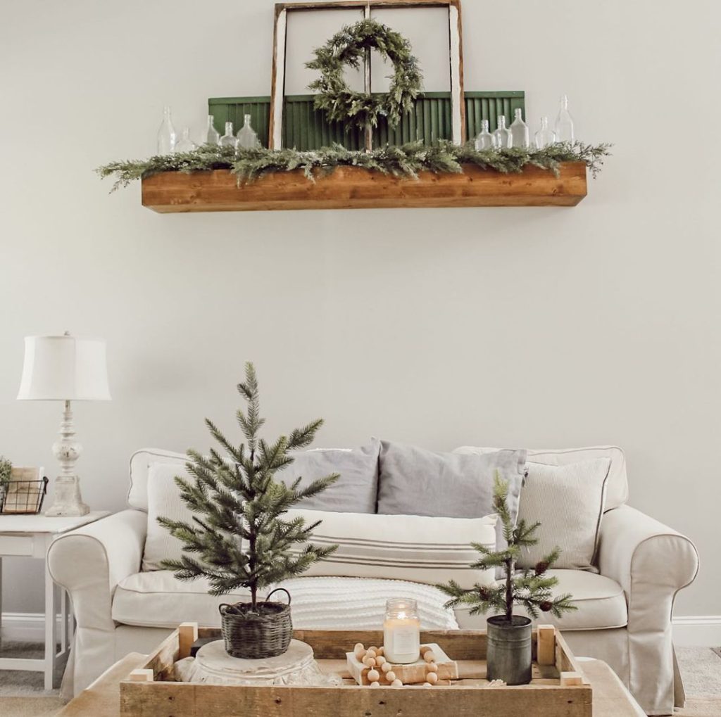 A cozy living room with a white sofa, a wooden coffee table adorned with small Christmas trees, a candle, and wooden beads. The wall shelf above features holiday greenery, a wreath, and glass decorations—perfect inspirations from 10 Cozy Winter Decor Ideas for a Chic Home.