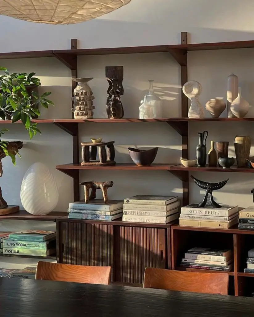 Wooden shelves displaying various sculptures, vases, and books add a touch of Beyond Books: 10 Creative and Stylish Uses for Your Bookshelves inspiration. A plant on the left and a large light fixture from above complete the look. In the foreground, a dark table with chairs invites you to sit and admire.