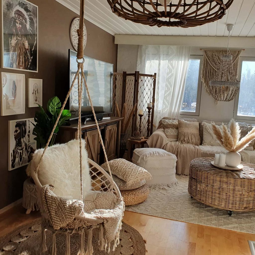 A cozy living room with natural, bohemian decor showcases how to curate your living room with Bohemian treasures. It features a hanging chair, a wicker coffee table, a sofa with cushions, and artworks on the wall. Large windows provide ample sunlight.
