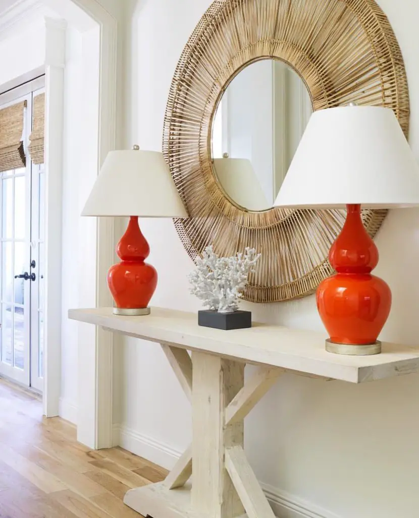 white entryway table with two red table lamps with white shades and a round wall mirror in wicker frame