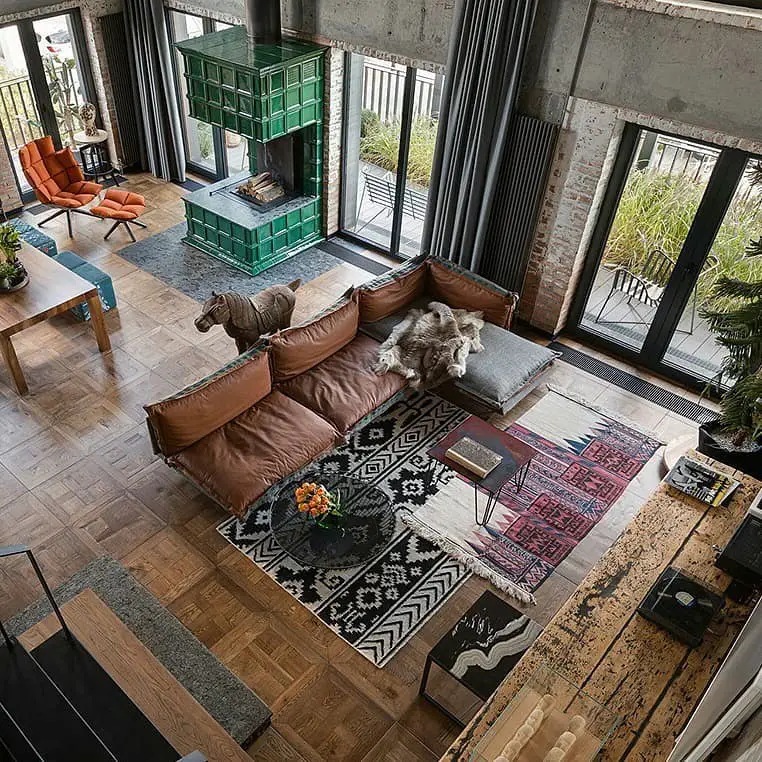 Spacious living area with wood and tile flooring, a large sectional sofa, patterned rugs, a green fireplace, and floor-to-ceiling windows. This inviting industrial living room haven is styled with various furniture and decor items arranged throughout the room.