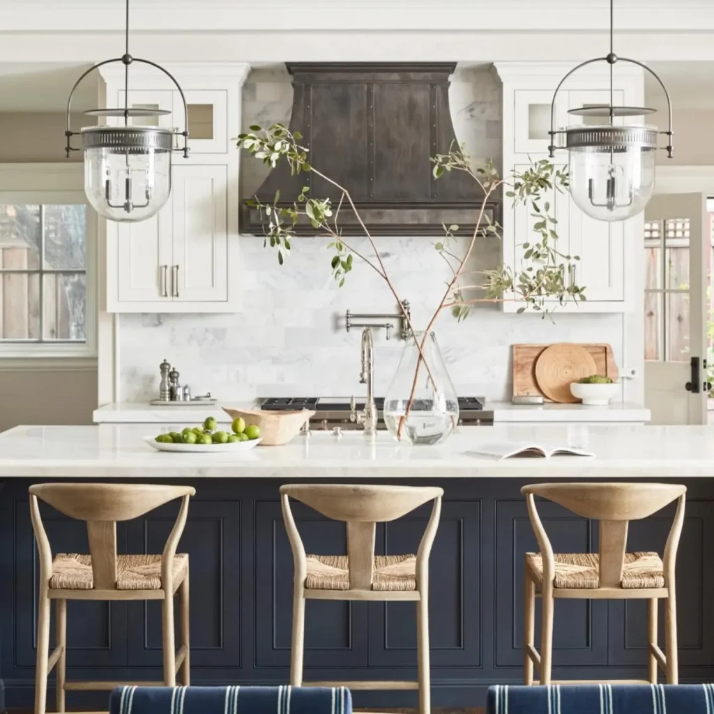 A modern kitchen with a large island features three wooden bar stools, white countertops, hanging pendant lights, a vase with branches, a dark hood over the stovetop, and subtle touches of coastal decor to achieve the modern coastal kitchen look.
