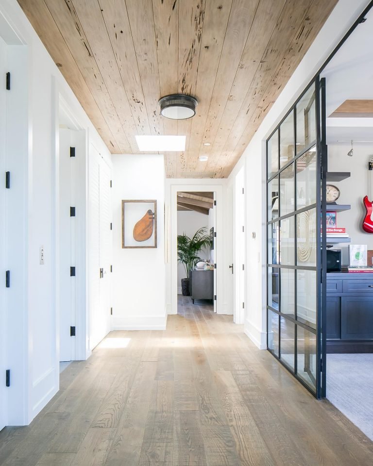 A hallway with wooden ceiling and floors, white walls, a door at the end, and a glass sliding door on the right. A painting and utility doors are on the left—perfect for exploring optical illusions that make rooms look bigger.