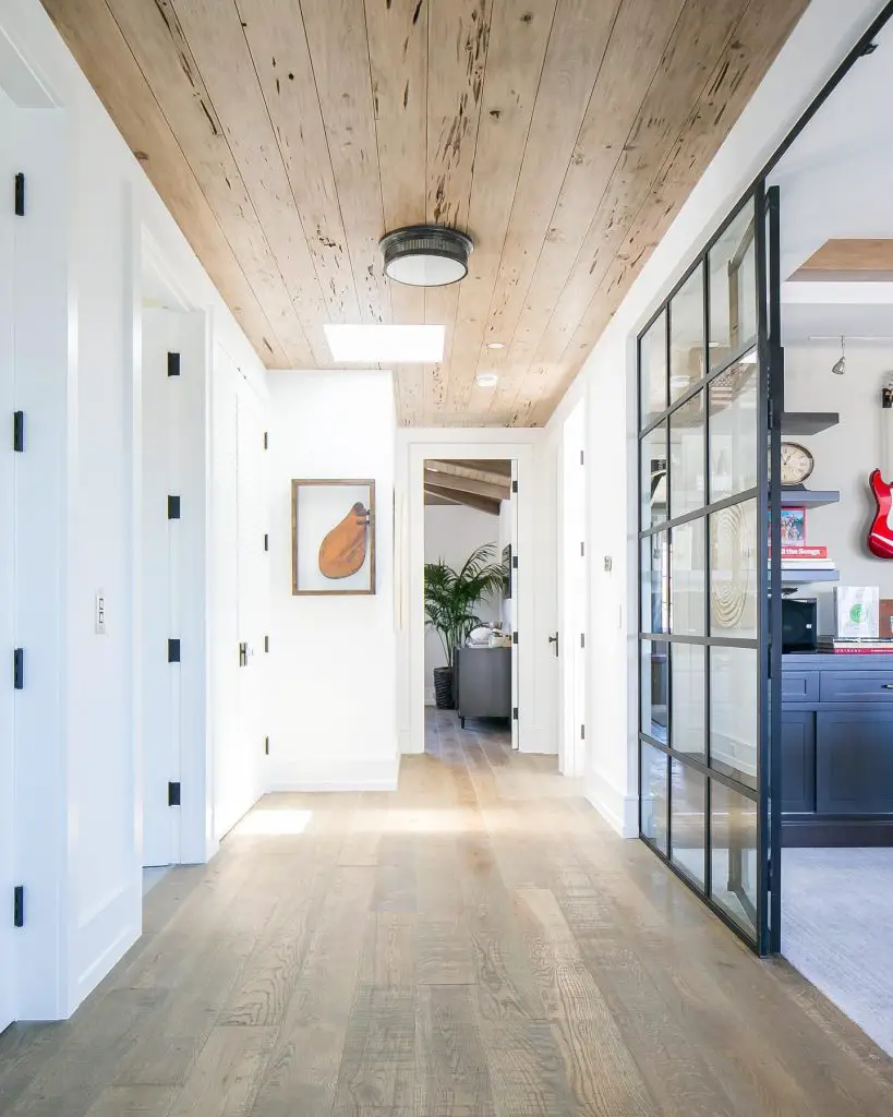 A hallway with wooden ceiling and floors, white walls, a door at the end, and a glass sliding door on the right. A painting and utility doors are on the left—perfect for exploring optical illusions that make rooms look bigger.