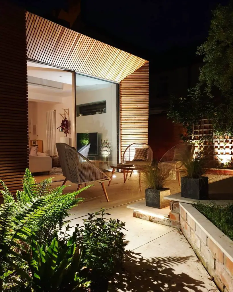 A nighttime view of a contemporary outdoor patio with wooden slats, chairs, plants, and can lights shaping the perfect outdoor mood, leading into a modern living space with a glass door.