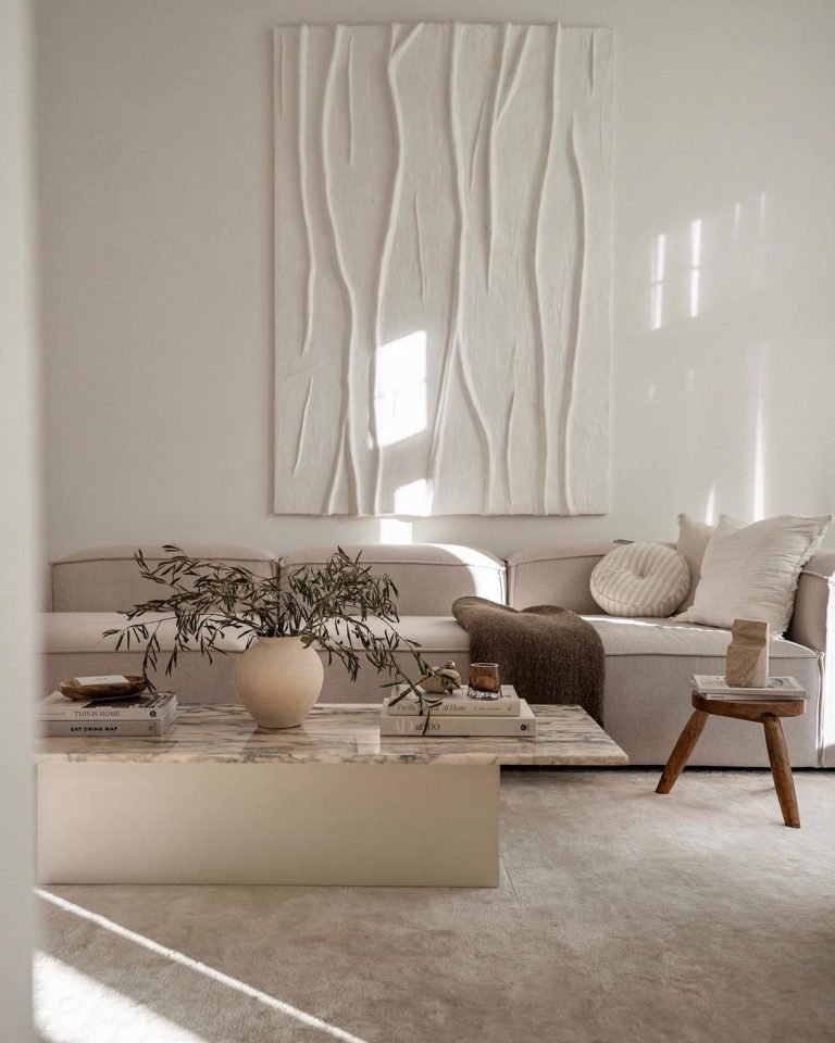 A minimalist Scandinavian living room features a white sectional sofa, a marble coffee table with potted plant and books, a wooden stool, and large textured wall art. Sunlight filters through the space, creating a serene ambiance perfect for crafting a stunning interior.