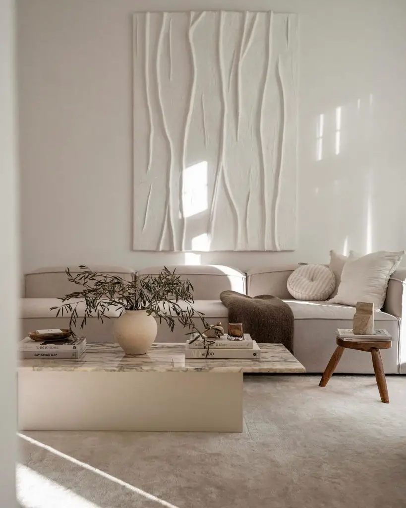 A minimalist Scandinavian living room features a white sectional sofa, a marble coffee table with potted plant and books, a wooden stool, and large textured wall art. Sunlight filters through the space, creating a serene ambiance perfect for crafting a stunning interior.