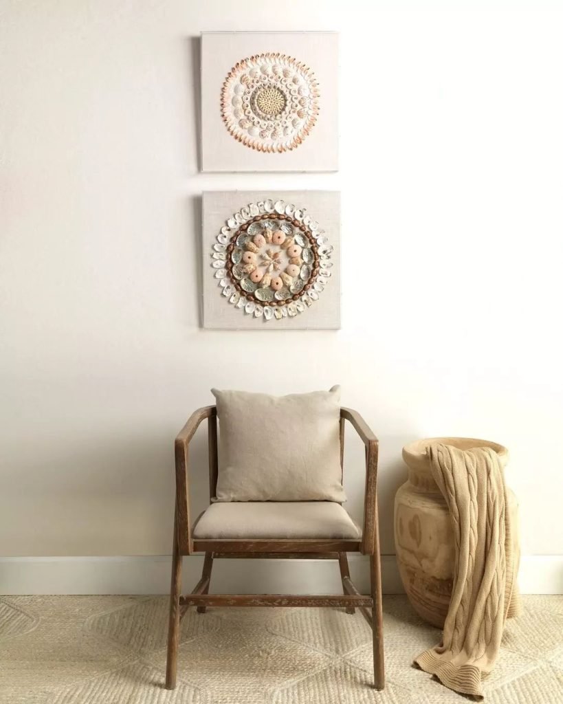 A wooden chair with a beige cushion, a decorative clay pot with a beige blanket, and two wall art pieces featuring mandala designs in neutral tones are arranged against a white wall. Consider incorporating seashell wall art ideas for a breathtaking display to elevate the coastal vibe.