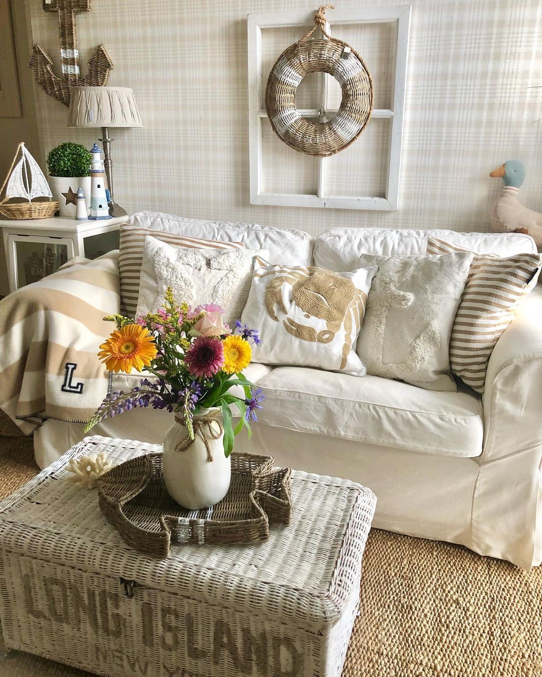 a farmhouse-coastal-style room with a couch with white slipcover, throw pillows, and a wicker storage box used as a coffee table styled with florals in a vase