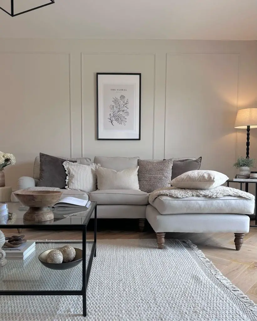 A neutral-toned living room showcases what it takes to master styling like a pro with throw pillows, featuring a beige sectional adorned with plush pillows, a glass coffee table with decor items, a framed botanical print above the sofa, and a floor lamp illuminating the space.