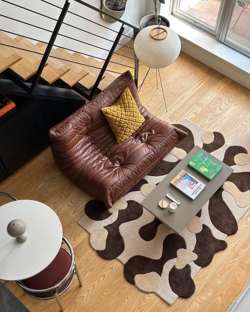 A modern living area with a brown leather sofa, a yellow cushion, and a patterned rug that shows what types of area rugs work best in small spaces. The room also features a coffee table with books and cups, a black railing staircase, and a standing lamp next to a large window.