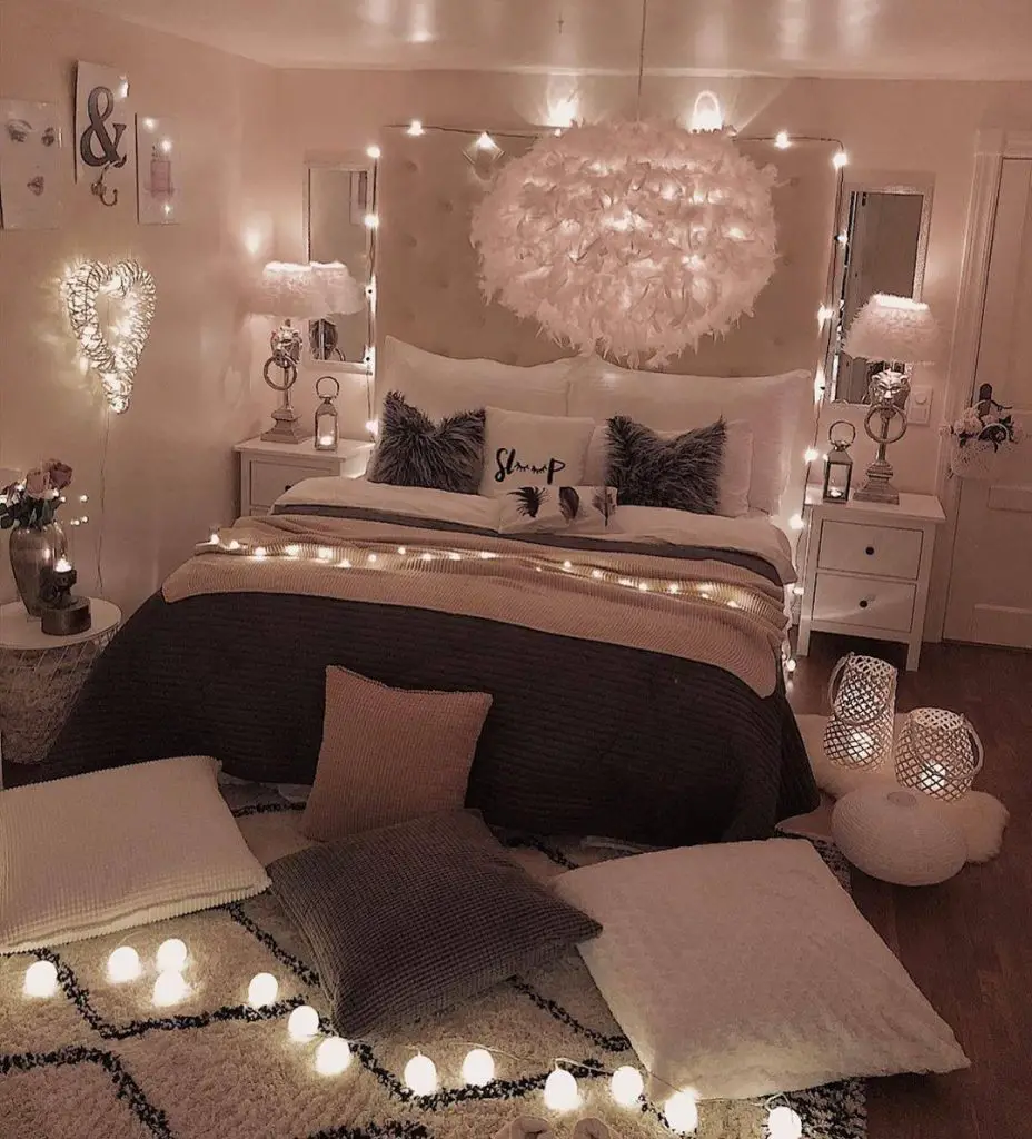 A cozy bedroom with a bed adorned with pillows and blankets, surrounded by fairy lights. There are cushions and small lights on the floor. Lamps and heart-shaped decorations are on the nightstands, creating a dreamy bedroom retreat.