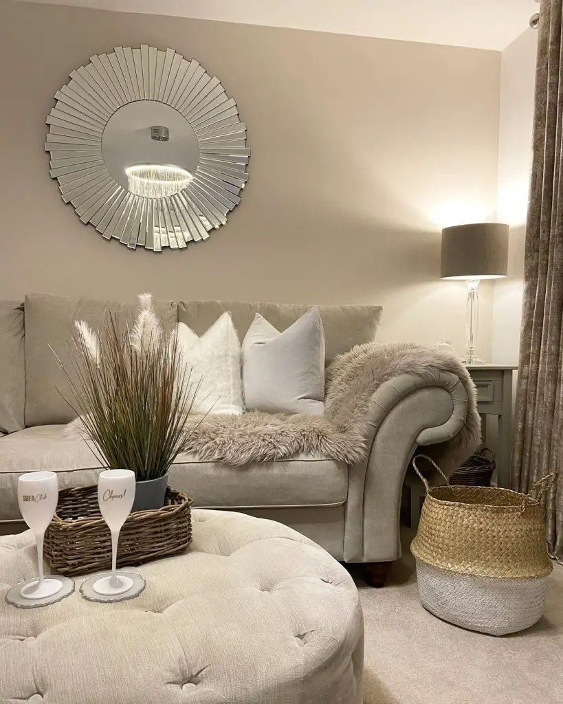 A cozy living room with a beige sofa adorned with white and beige cushions, a decorative mirror on the wall above, a round ottoman, and a table lamp. Pro tips for a warm and stylish winter decor: add faux fur throws or pillows for extra warmth. A basket and two wine glasses on a tray complete the look.