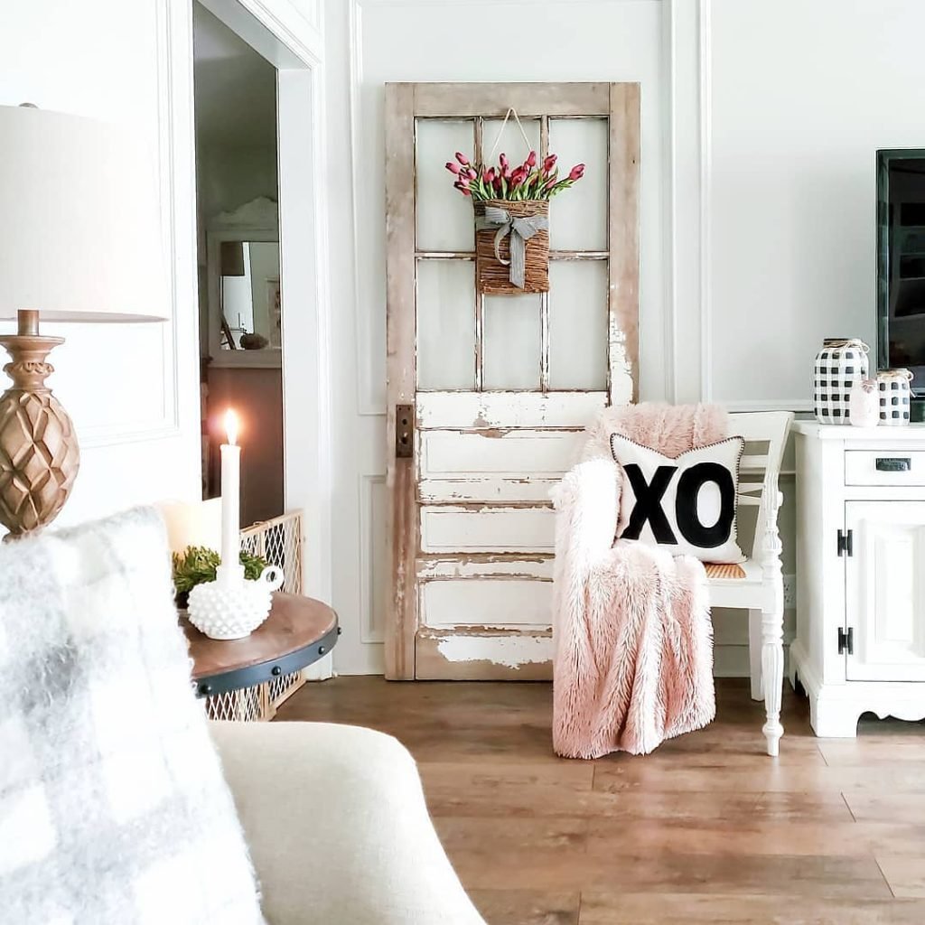 A cozy living room with a rustic wooden door adorned with a basket of flowers. The space features a candlelit table, a white cabinet, and a chair with a pink throw and XO pillow. For an extra touch of warmth this winter, try adding a faux fur throw or pillow to complete the look.