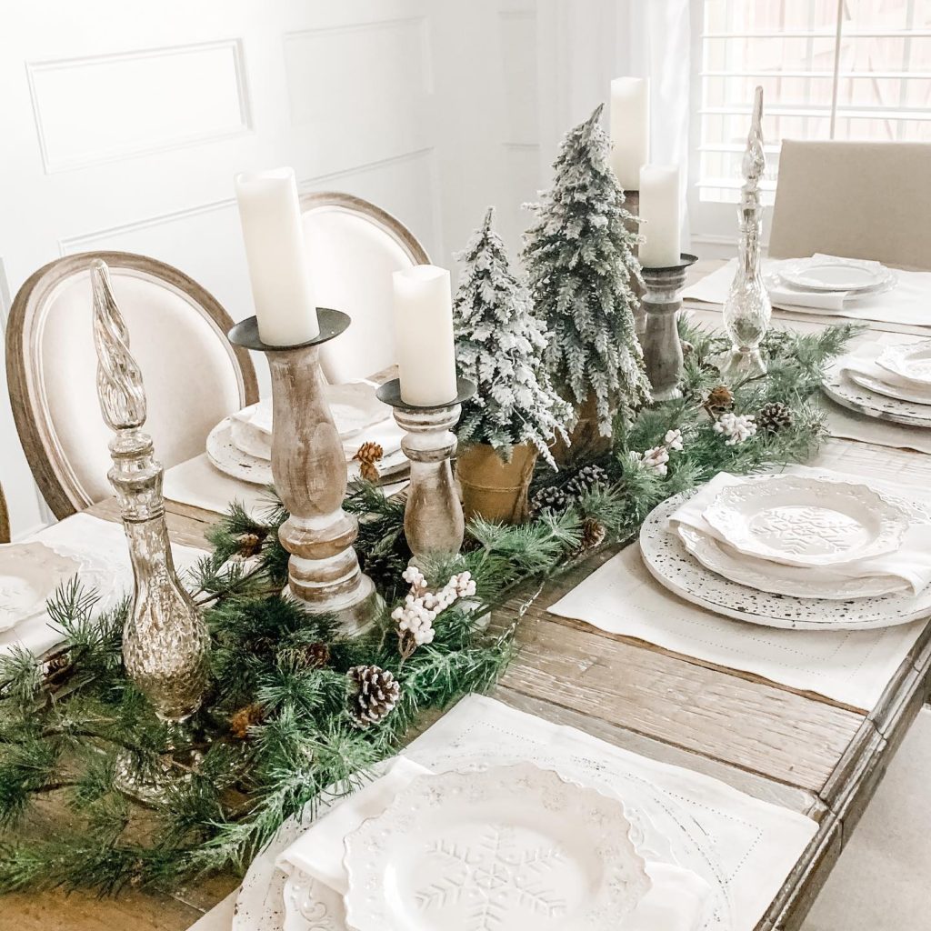 A dining table set for six with white plates, silverware, candles, and a centerpiece featuring artificial snow-covered trees and pine garland evokes Minimalist Magic: How to Style a Simplified Winter Table Decor.