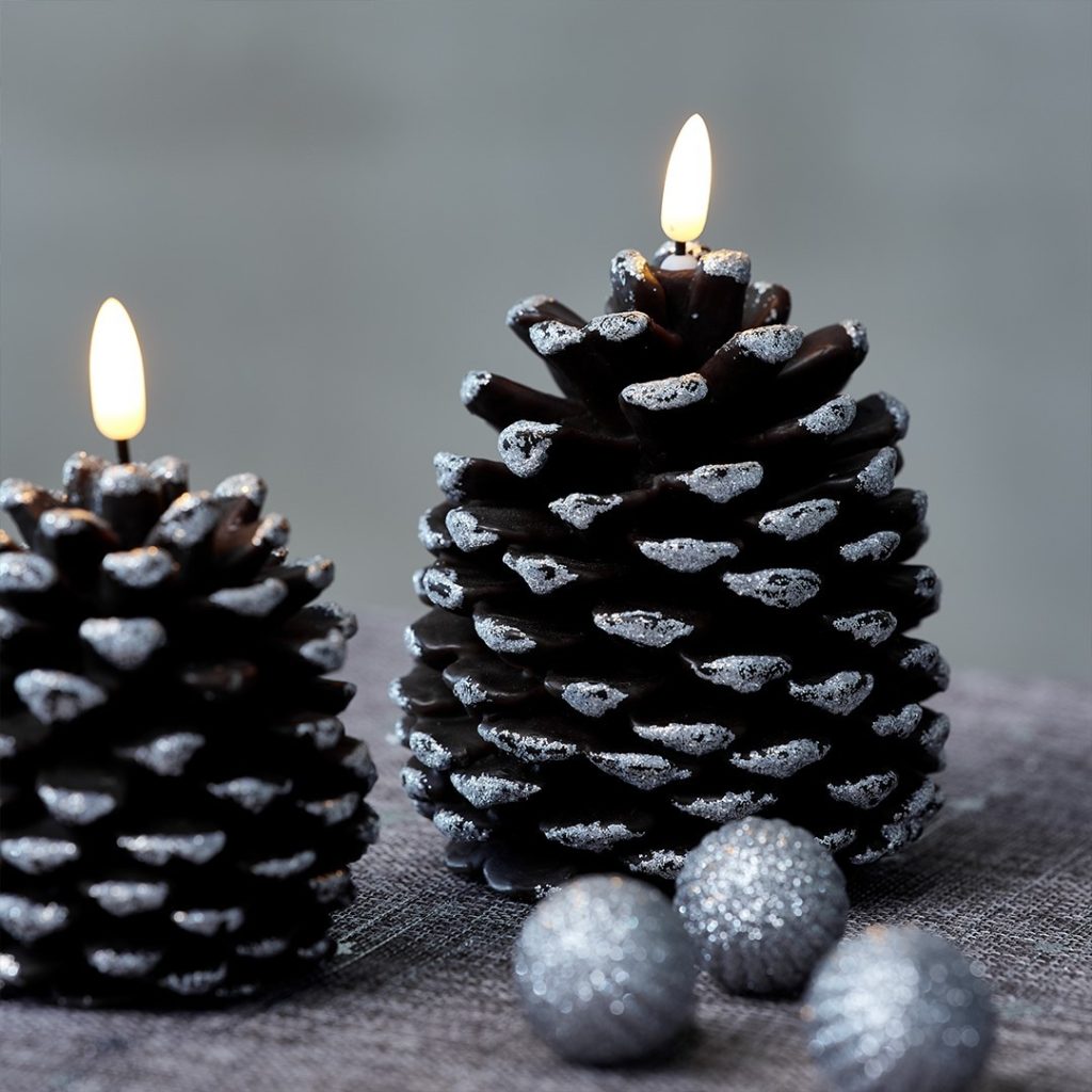 Two lit pinecone-shaped candles with glittery tips are placed on a surface next to three small glittery silver balls, creating Pinecone Magic: Transforming Your Home for a Festive Christmas.