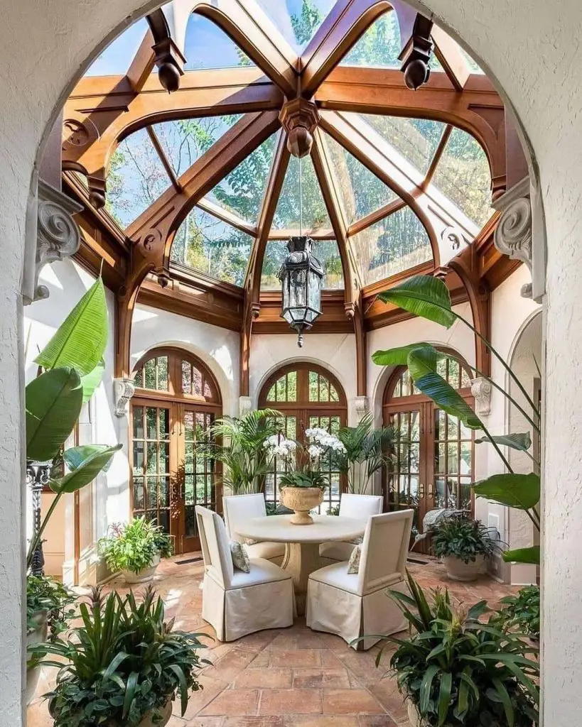 A sunlit conservatory with wooden beams, large windows, and a glass ceiling, featuring a round table with four chairs, surrounded by plants. Discover how you can create soothing spaces through greenery in this serene setting.