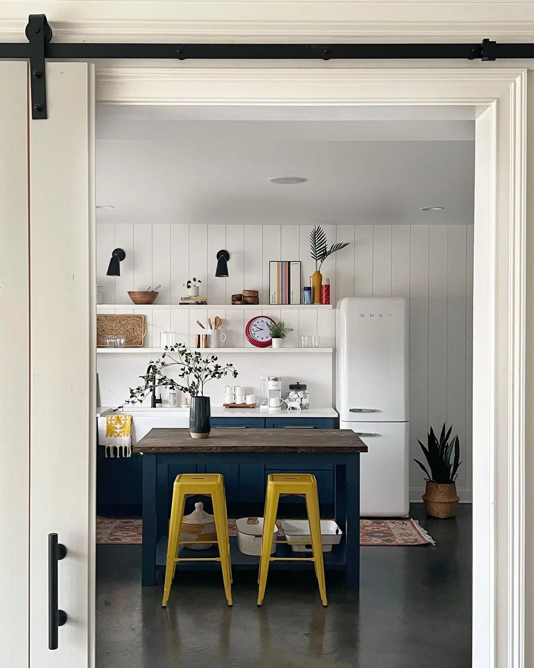 A modern kitchen with white walls, a wooden island with two yellow stools, open shelves with various decor, and a Smeg refrigerator that adds retro style.