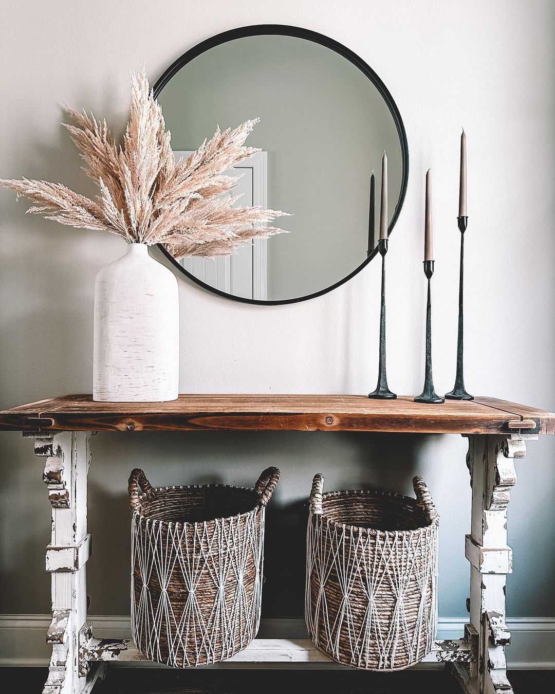 entryway console styled with a flower vase and candles holders with storage baskets underneath and round wall mirror