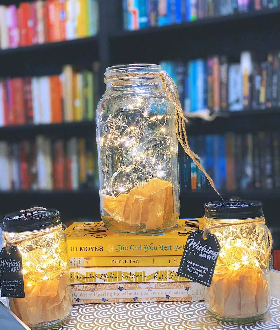 Three glass jars filled with string lights are arranged on stacked books with a bookshelf in the background. Two small jars have "Wishing Jar" labels, adding a touch of Mason Jar Magic: Lighting Up Your Holidays with Festive Flair.