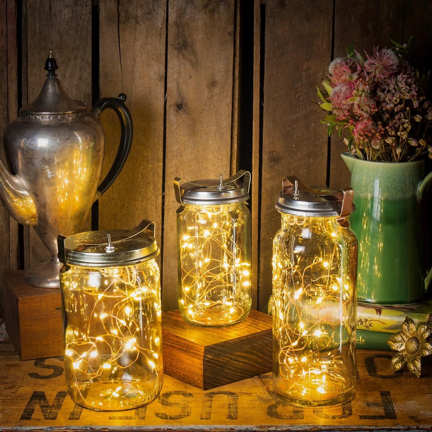Mason Jar Magic comes to life as three mason jars filled with string lights sit on wooden boxes next to a pewter pitcher and a green vase with flowers, all set against a rustic wooden background, lighting up your holiday decor with festive flair.