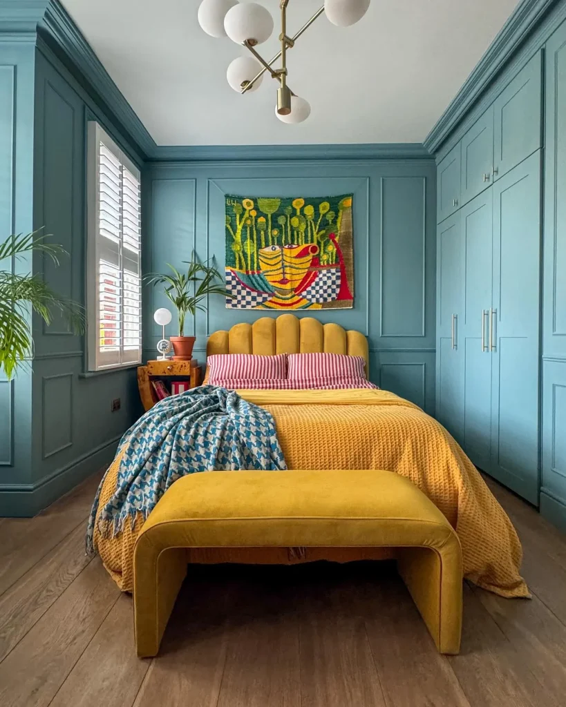 A bedroom with blue paneled walls, a yellow upholstered bed and bench, striped pillows, a colorful wall hanging, and plants by the windows illustrates how to create balance in eclectic decor: fundamental principles bring harmony to diverse elements.