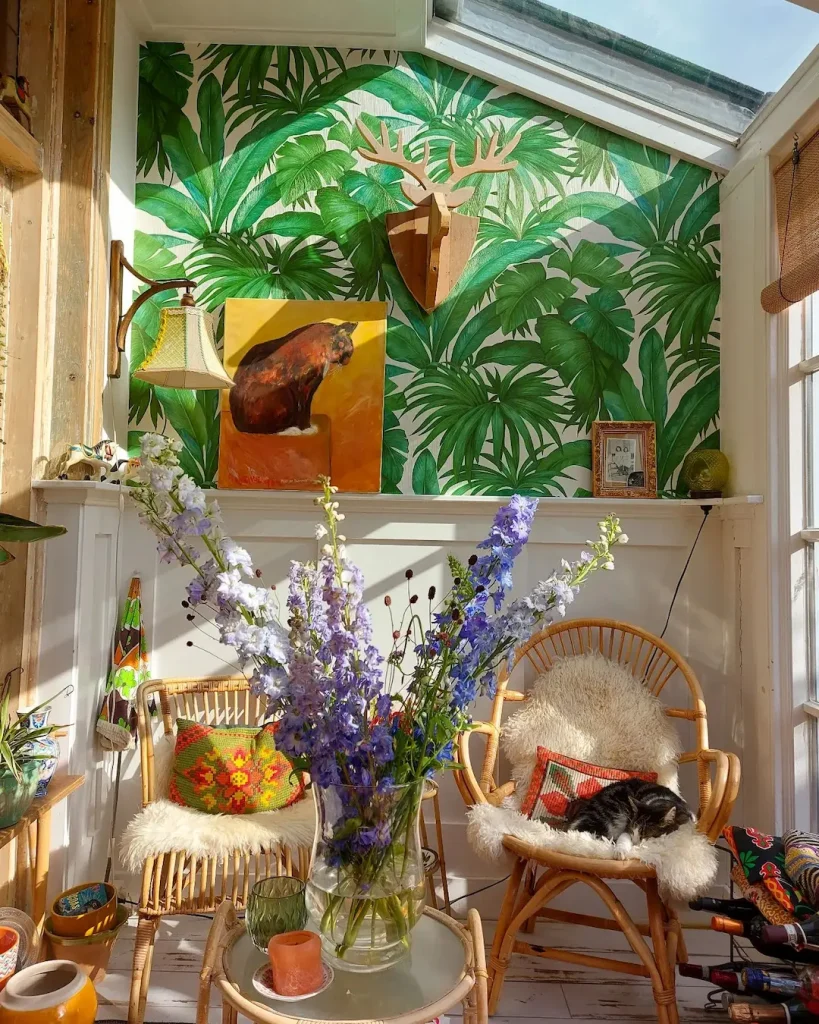 A sunlit room with wicker chairs, fluffy cushions, and a vase of purple flowers captures the essence of funky bohemian living room décor. With green leafy wallpaper adorning the walls, a wooden deer head and a bear painting add unique charm—complemented by a peacefully sleeping cat.