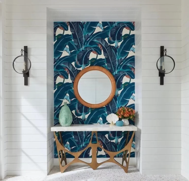 A funky console gives this entryway with contrasting bright blue palm wallpaper against stark white walls along with a rattan mirror and two wall sconces an unexpected but nice addition to create an inviting and striking entryway decor.