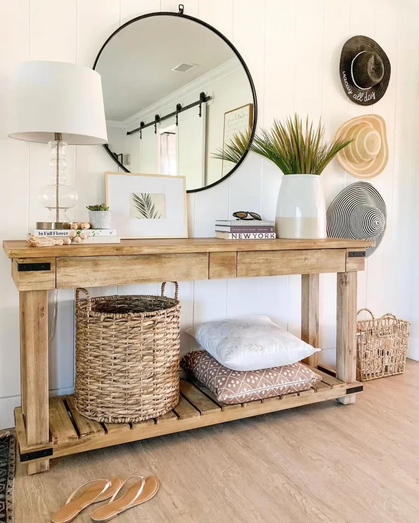 what's not to love in this summer entryway decor that includes a lovely wooden console table styled with a beautiful vase, books, lamp, framed picture, pillows, and a basket while the wall is decorated with a round mirror and a hat wall!