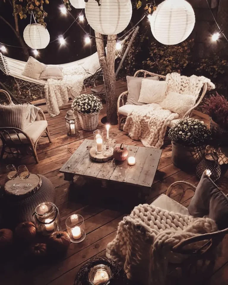 Cozy outdoor patio at night with string lights, lanterns, and candles. Cushioned seating around a wooden table, a hammock in the background, and various blankets and plants add to the ambiance. Discover how to style your cozy bohemian patio with vintage furniture for an inviting atmosphere.