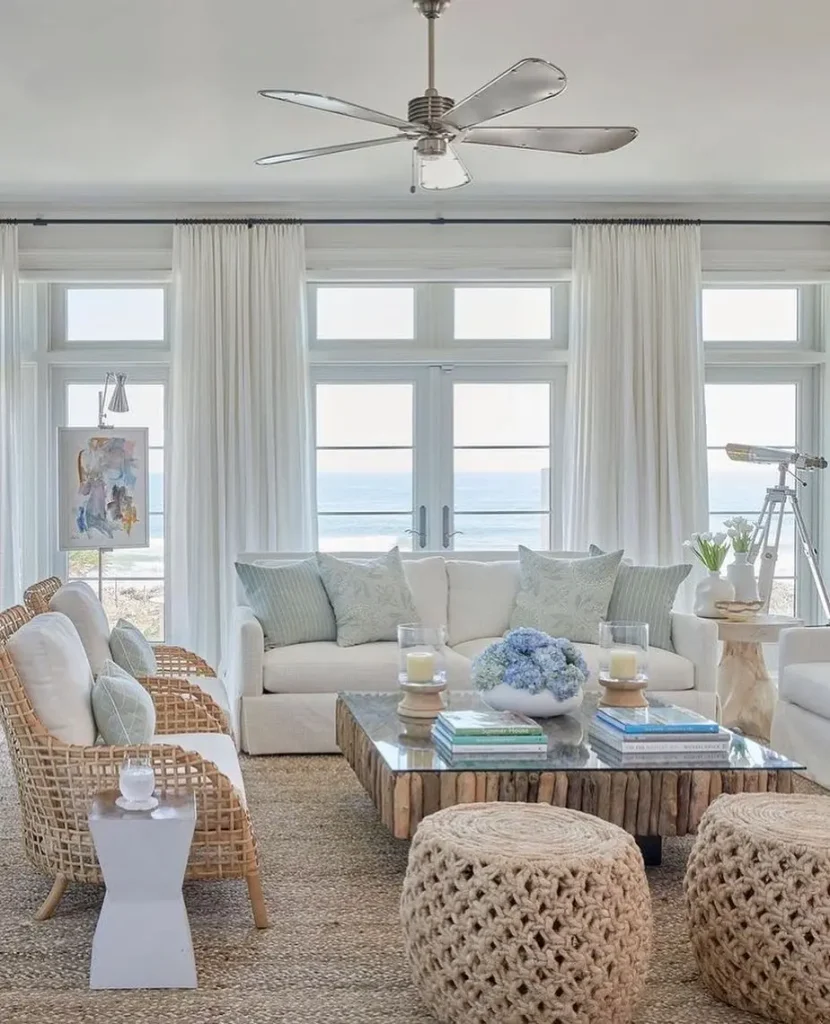 A bright living room with a white sofa, wicker chairs, and a glass coffee table highlights how to design a beach-styled living room on a budget. Large windows with sheer curtains offer an ocean view, complemented by a ceiling fan and telescope for relaxation.