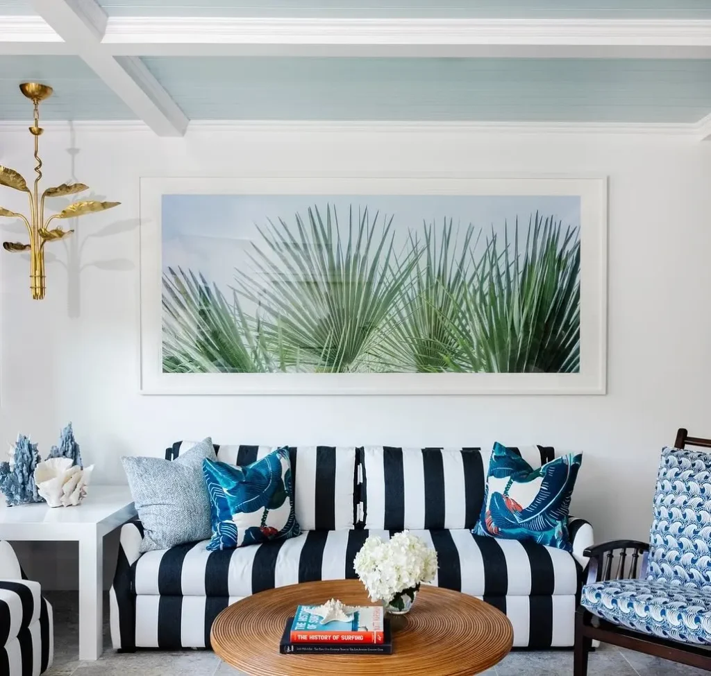 Living room with a large framed photo of palm leaves above a black and white striped sofa, blue-patterned pillows, a round wooden table, a stack of books, and a flower arrangement—perfectly showing one of the 5 Coastal Color Palettes: How to Refresh Your Home.